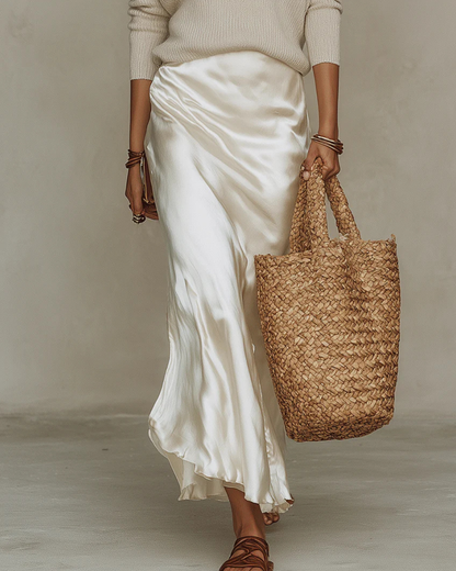 Lower-body view of the flowing cream satin midi skirt with softly draped folds, worn with strappy brown sandals and a straw tote. 10