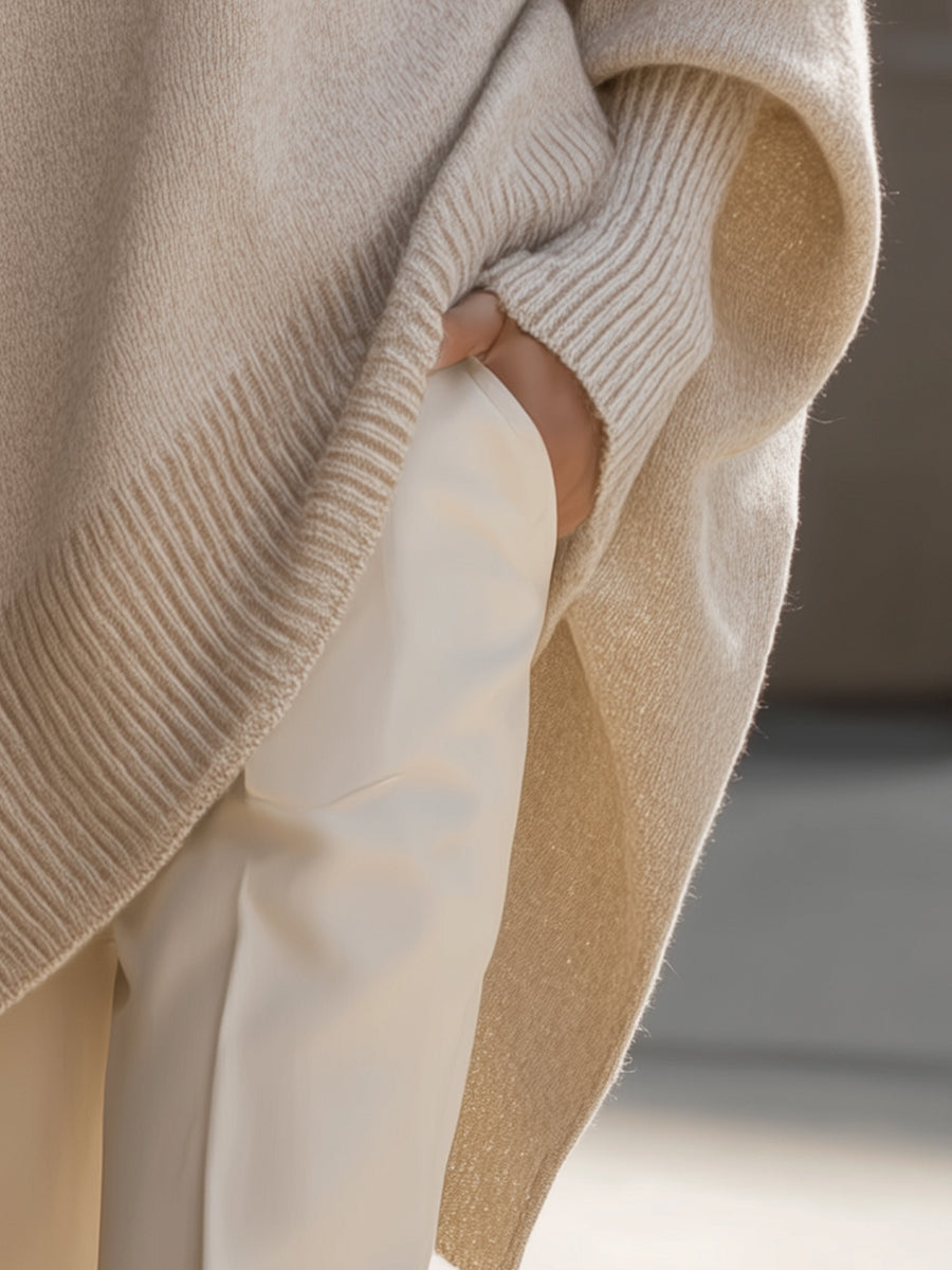 Detail of the beige poncho's side slit and ribbed hem, with a hand resting in a trouser pocket 6