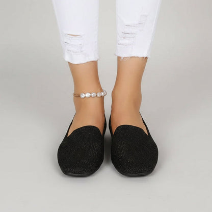 Front view of black sparkly knit slip-ons worn with white distressed jeans and a pearl anklet against a plain grey background. 2