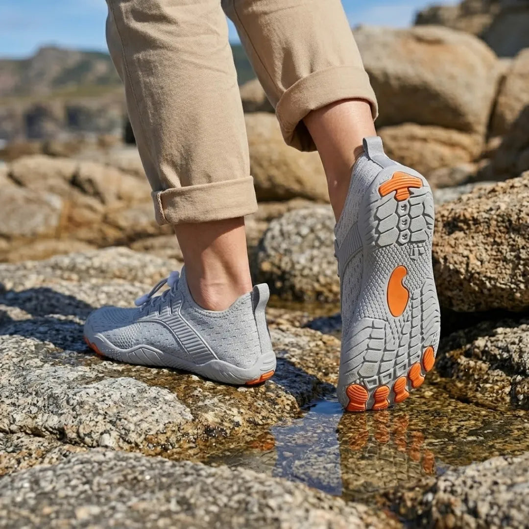 Grey barefoot trainers worn on wet coastal rocks, one sole raised to show grey tread with orange toe pads and centre patch. 11
