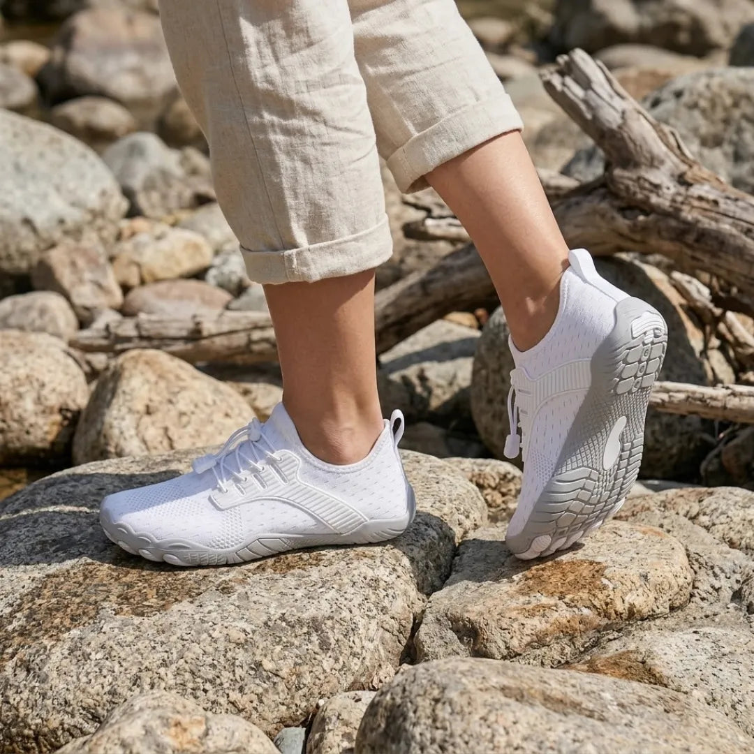 White barefoot trainers worn on sunlit rocks, one foot raised to show the grey patterned sole, with rolled linen trousers. 2