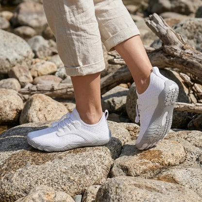 White barefoot trainers worn on sunlit rocks, one foot raised to show the grey patterned sole, with rolled linen trousers. 2