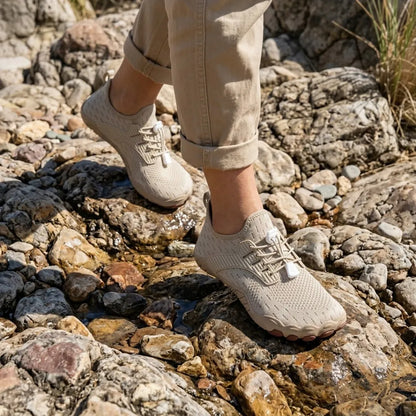 Beige barefoot trainers worn while stepping across wet, rocky ground, with cuffed trousers and laces fastened with white toggles. 20
