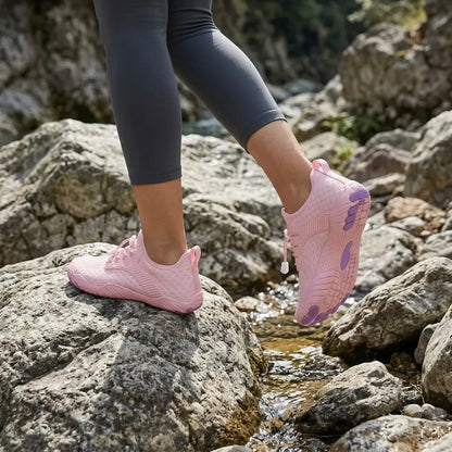 Woman in grey leggings stepping across rocks by a stream in pink barefoot trainers with purple toe pads. 29
