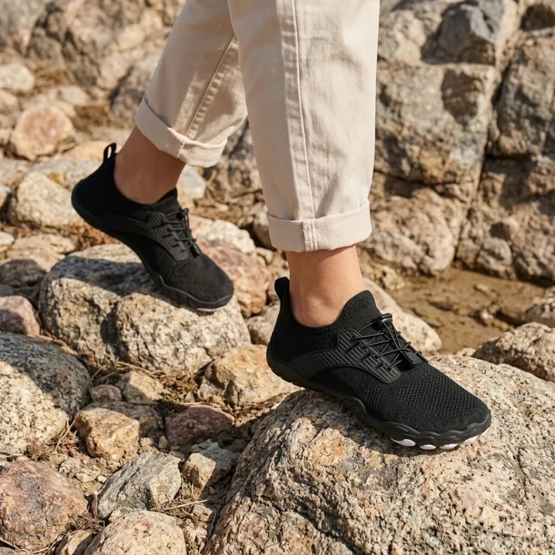Person in rolled-up cream trousers wearing black barefoot trainers while walking over sunlit rocks. 38