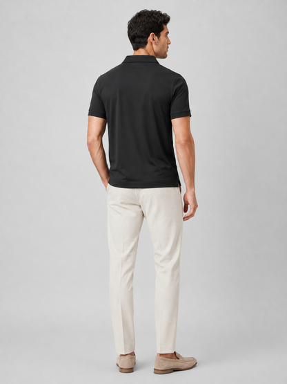 Back view of the black polo on a standing model, showing the plain back, short sleeves and small side vent, with cream trousers. 9