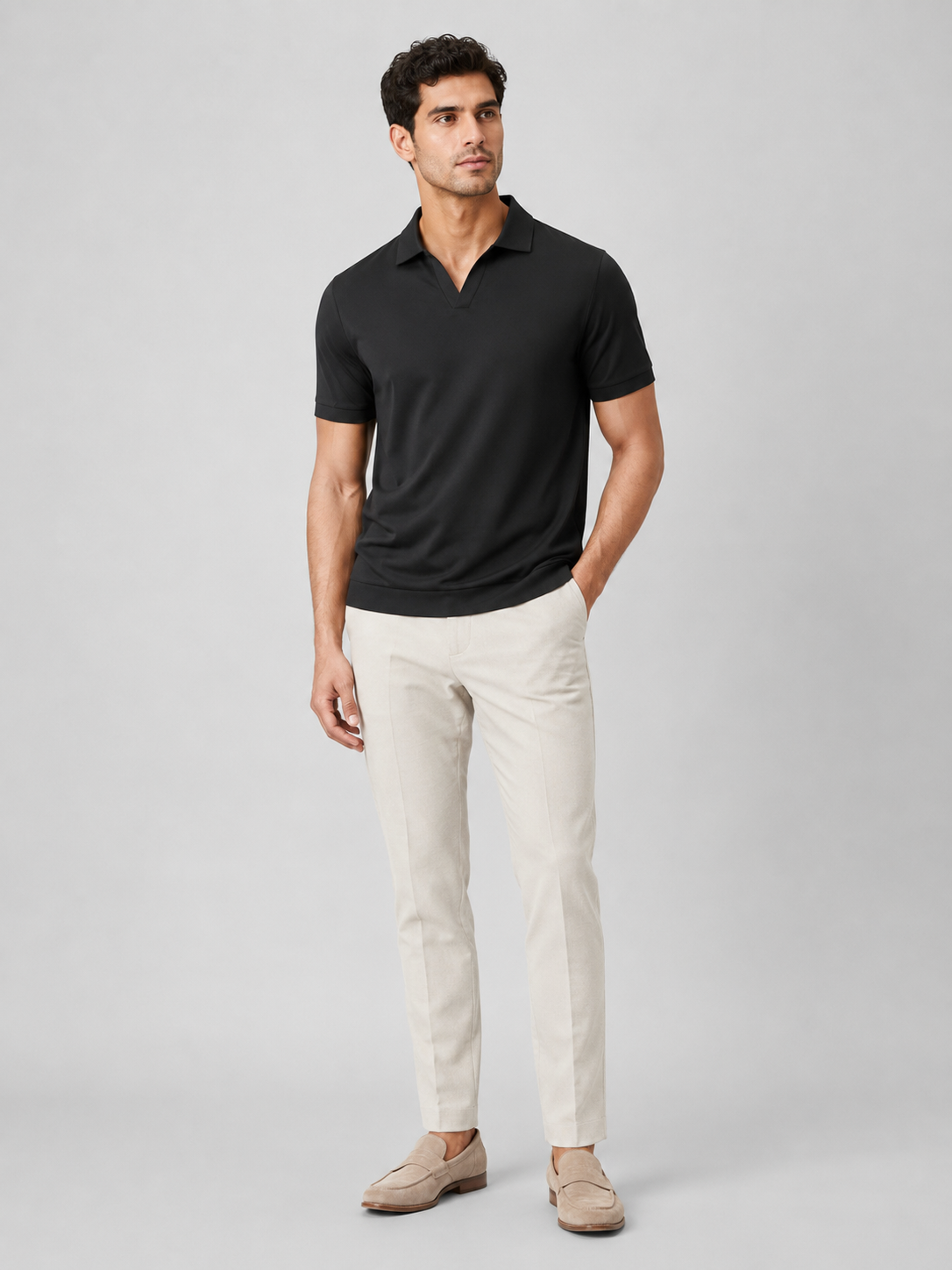 Full-length studio view of the black polo worn with cream trousers and tan suede loafers, one hand in pocket. 8