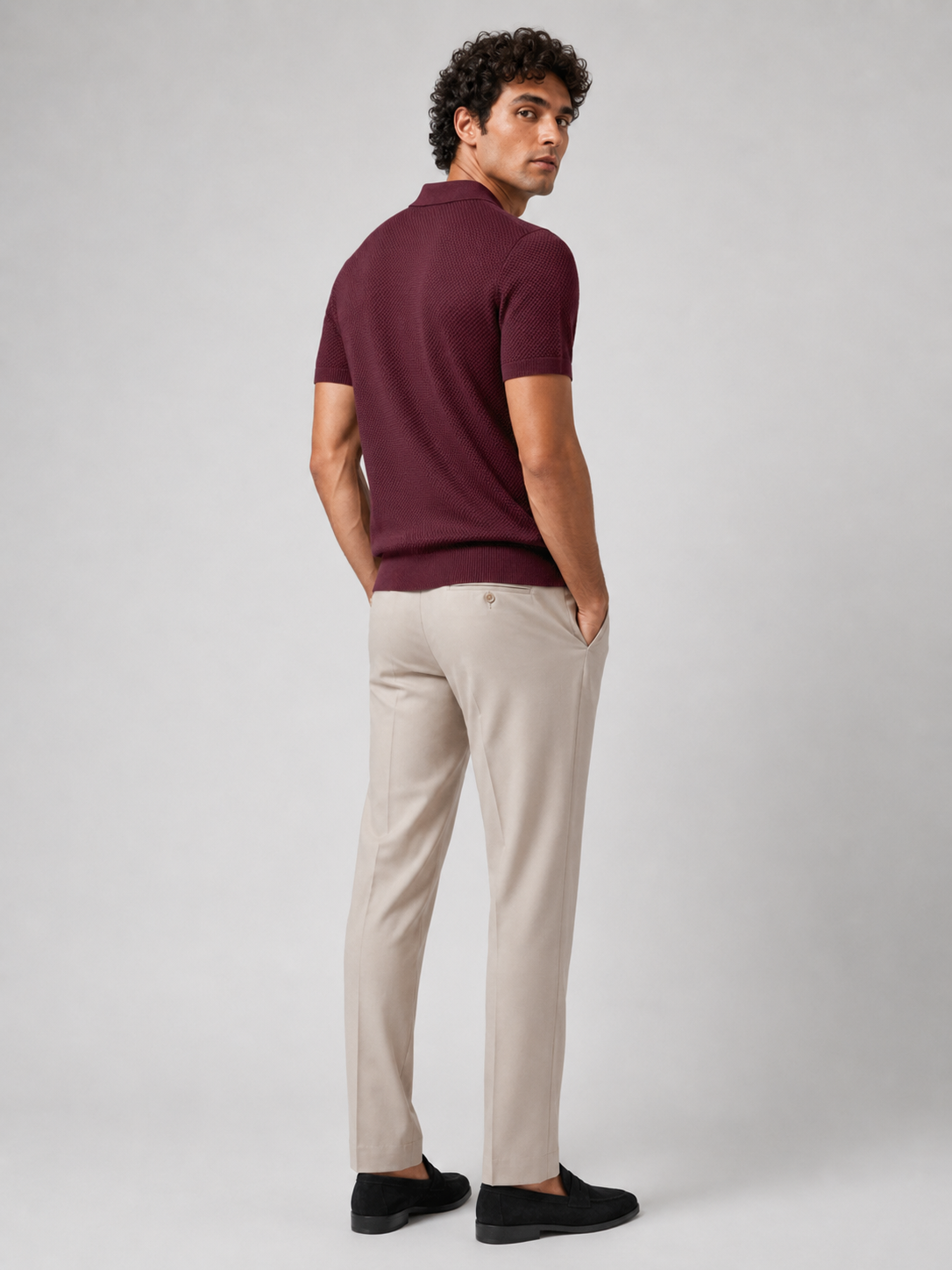 Back view of burgundy knit polo on model glancing over shoulder, with beige trousers and black loafers 24