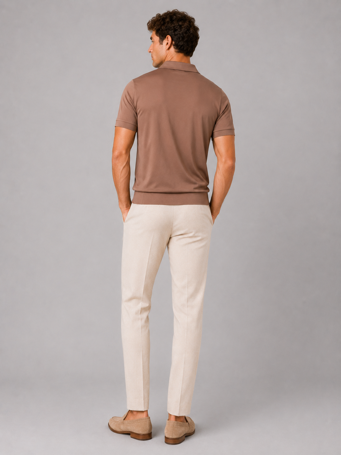 Back view of the brown polo on a standing model, showing the plain back, ribbed hem and short sleeves, with cream trousers. 14