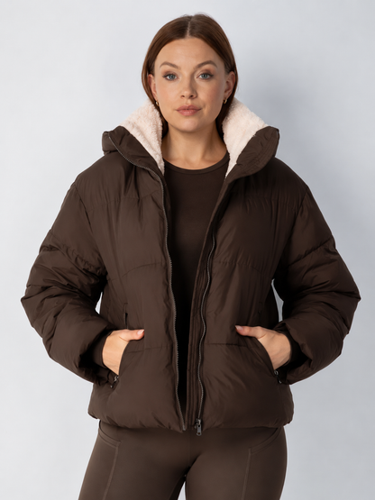 Model in a dark brown puffer jacket worn open with cream fleece lining at the collar and hands in the pockets 14