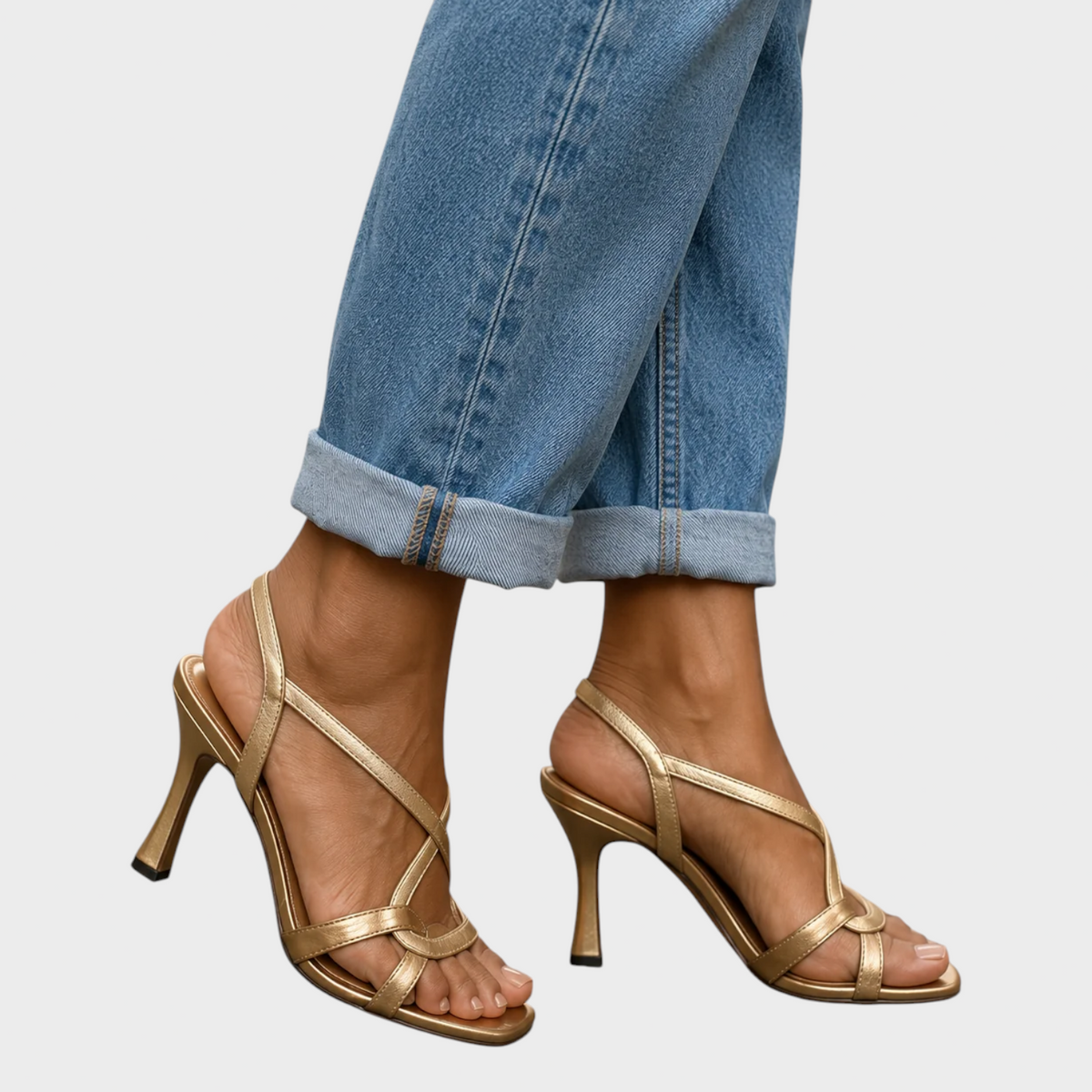 Side view of a model walking in gold slingback sandals with thin crossed straps and flared high heels, beneath cuffed denim. 2