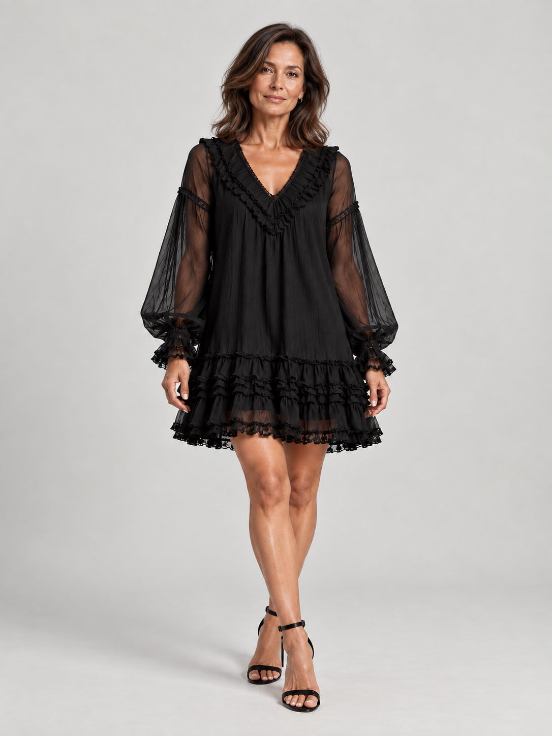 Full-length front view of a model in the black ruffled mini dress with sheer sleeves, styled with black ankle-strap heels. 2