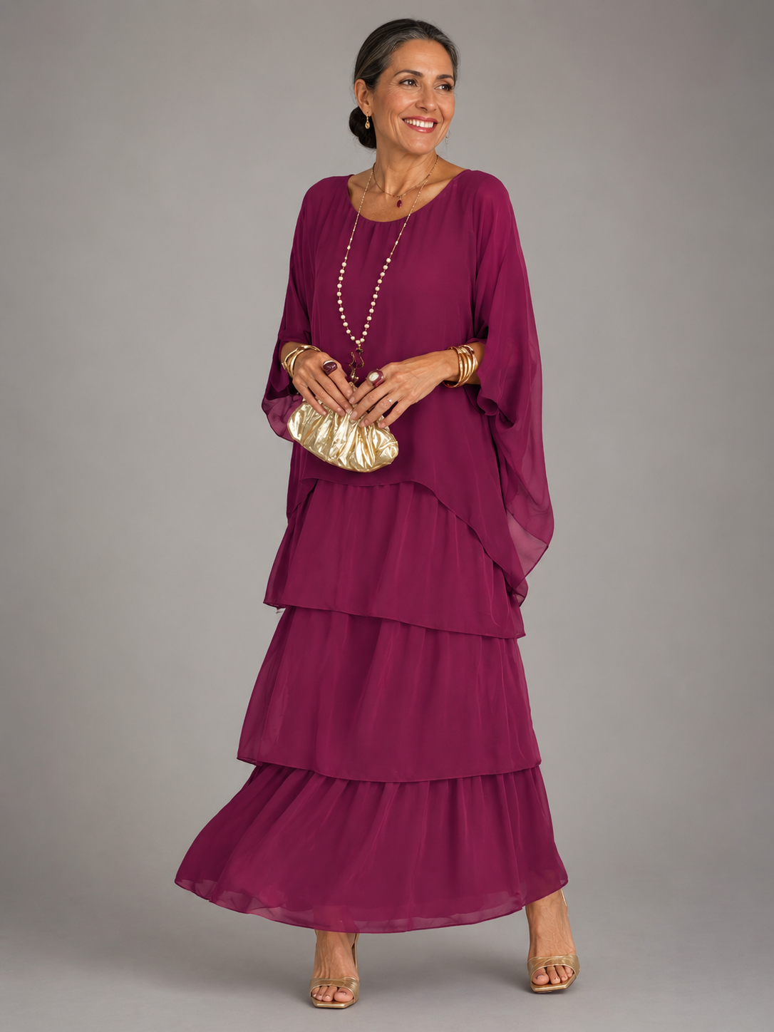 Woman in the deep magenta tiered chiffon maxi dress with batwing cape sleeves, gold bracelets and a gold clutch bag. 4