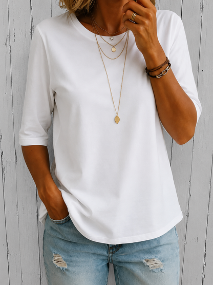 Woman wearing the round-neck half-sleeve t-shirt in white, with layered gold necklaces, brown bracelets and ripped light jeans, against weathered grey boards. 2
