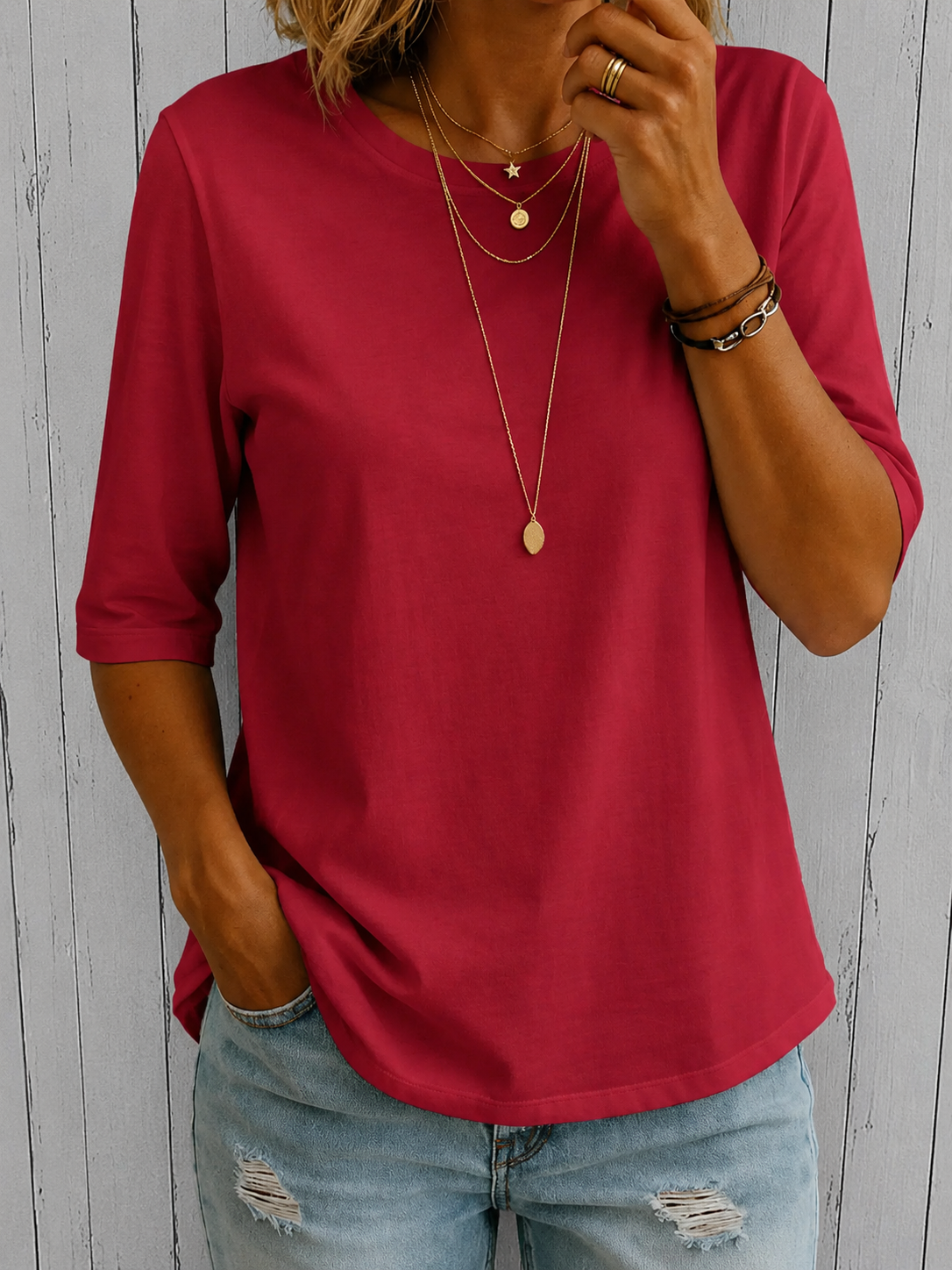 Model wearing the tee in deep raspberry red with layered gold necklaces and ripped light jeans, against pale wooden boards. 8
