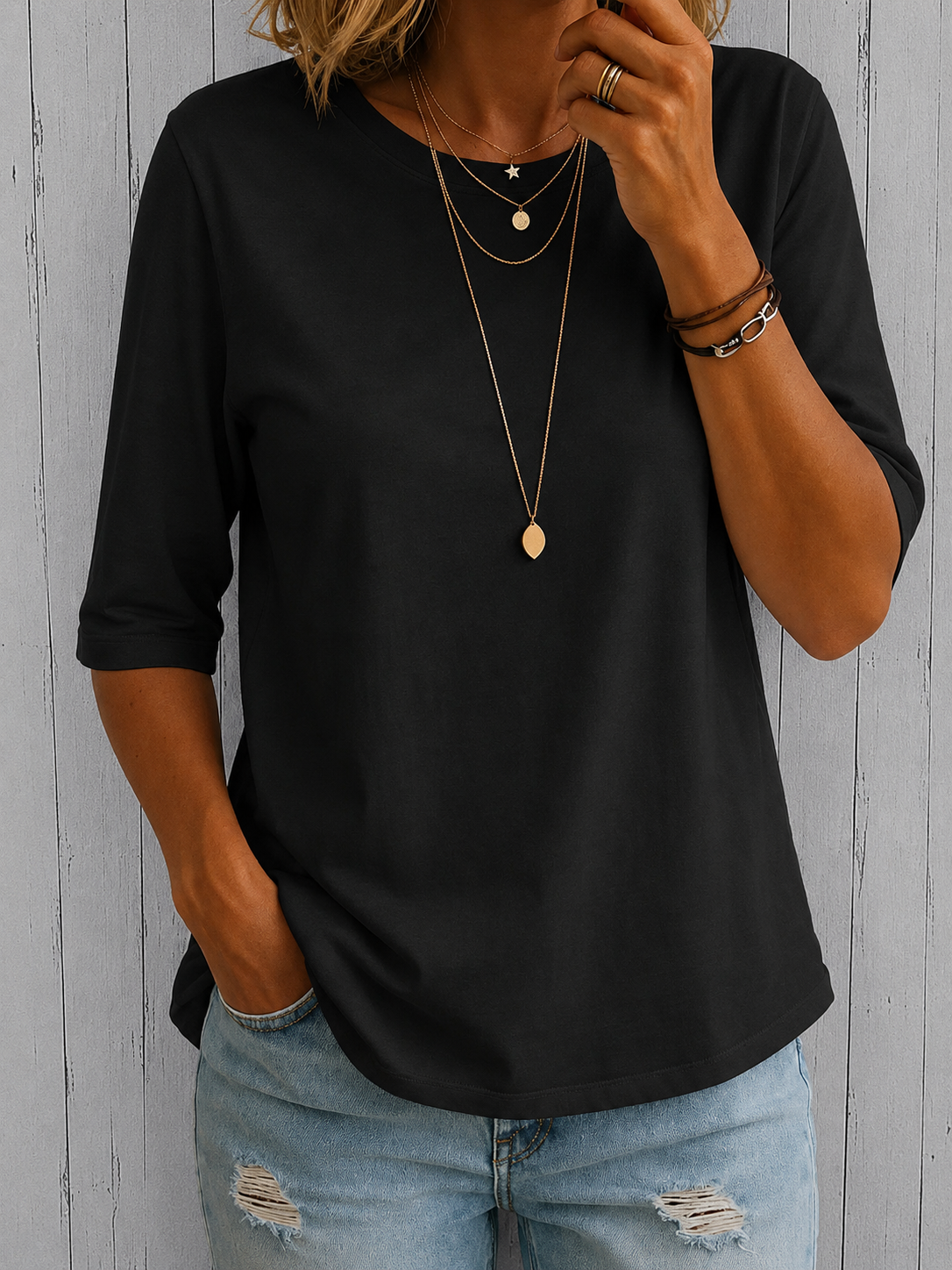 Model wearing the tee in black with layered gold pendants and ripped light-wash jeans, against grey wooden boards. 4