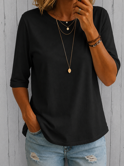 Model wearing the tee in black with layered gold pendants and ripped light-wash jeans, against grey wooden boards. 4