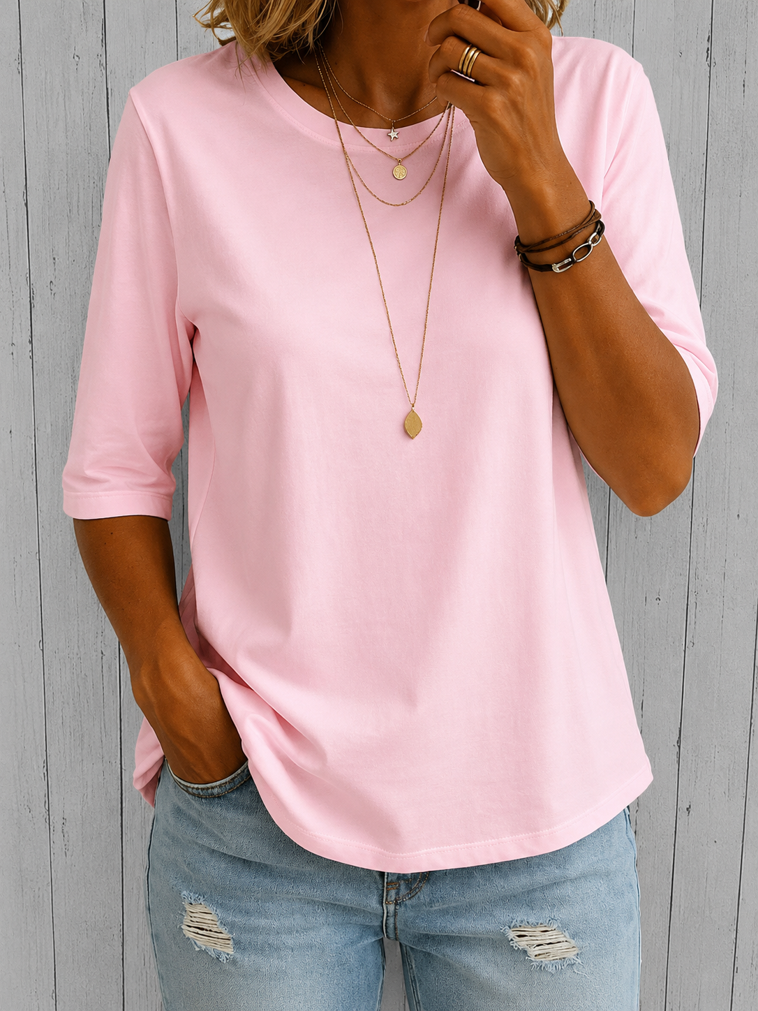 Model wearing the tee in soft pink with layered gold necklaces and leather bracelets, with a hand tucked into ripped jeans. 5