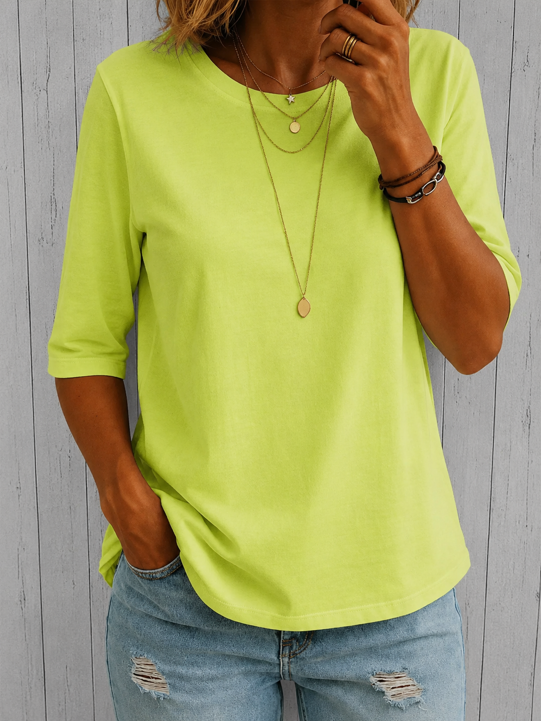 Model wearing the tee in bright lime green with gold necklaces and ripped jeans, standing against grey wooden panelling. 6