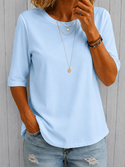 Model wearing the tee in pale sky blue with gold necklaces and ripped light jeans, standing before grey wooden panelling. 3