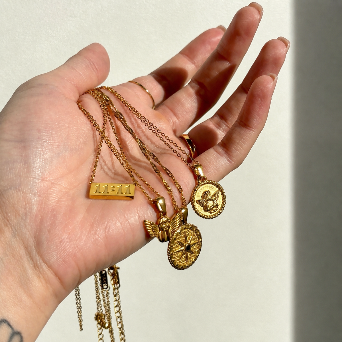 Open hand holding several gold pendant necklaces, including the round cherub medallion, a star disc, a small winged figure and a number bar. 3