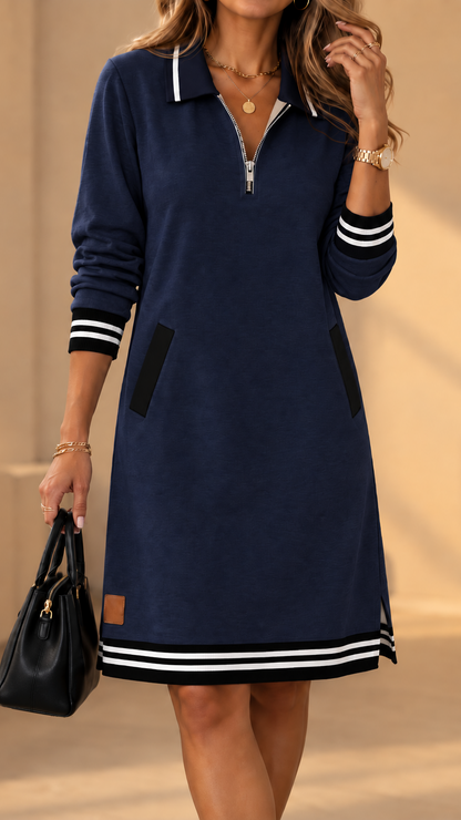 Model wearing the dress in navy blue with black and white striped cuffs and hem, front zip and side pockets, holding a black handbag. 2