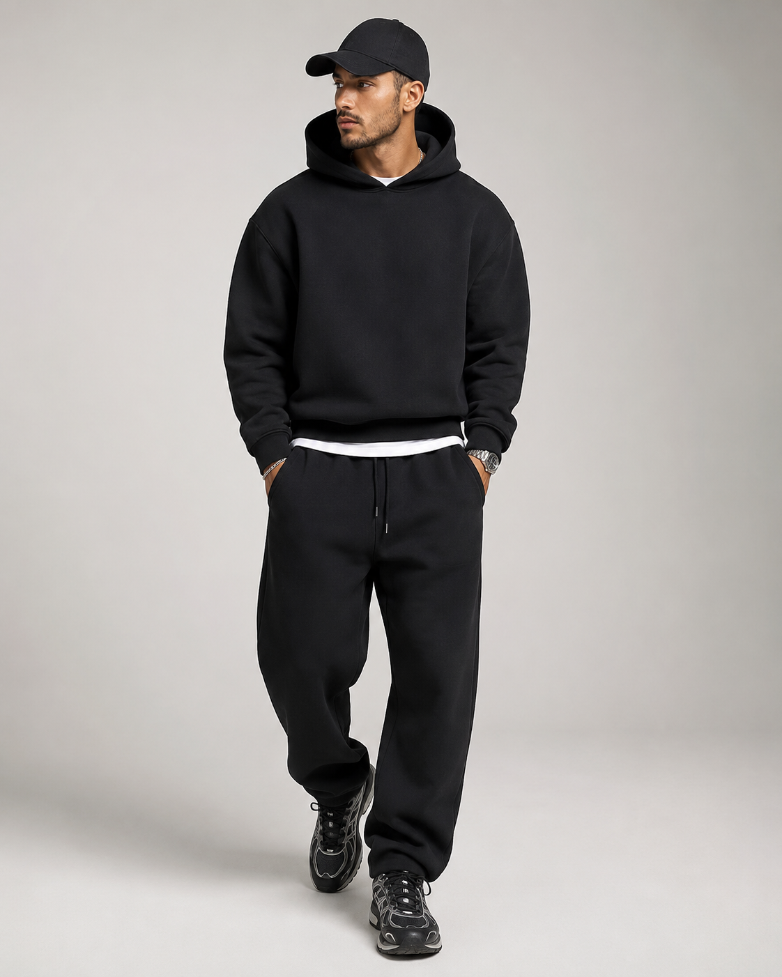 Model in the black hoodie and joggers walking, wearing a black cap, white layer and chunky black trainers, looking to one side. 7