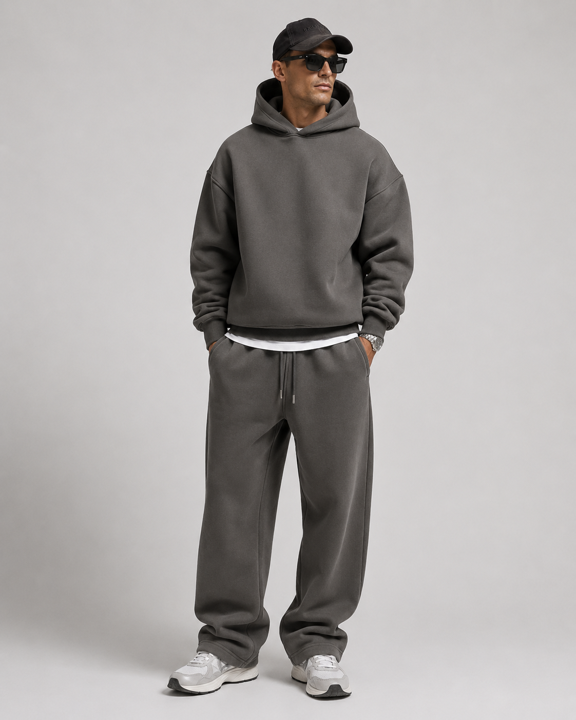 Studio shot of a model in the charcoal grey hoodie and relaxed joggers, with a dark cap, sunglasses and white trainers. 9