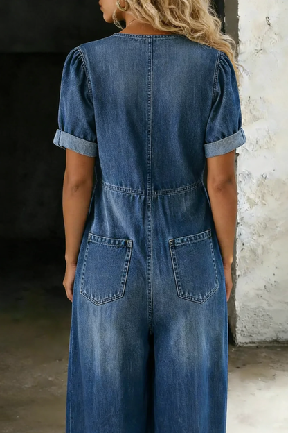 Back view of the mid-blue denim jumpsuit worn by a model, showing two back pockets and cuffed short sleeves 5