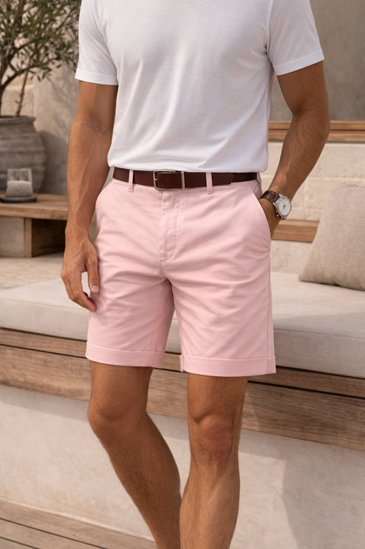 Front view on a model of pink turn-up shorts with a white T-shirt, brown leather belt and watch, one hand in pocket. 1