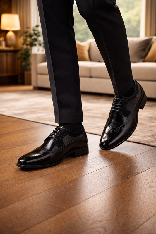 Glossy black leather wingtip brogues worn by a model mid-step in dark trousers, on a wooden floor in a living room. 2
