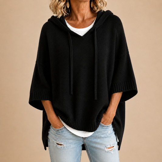 Black knitted hooded poncho with drawstrings and ribbed cuffs, worn over a white top and ripped light jeans, close-up of torso. 1