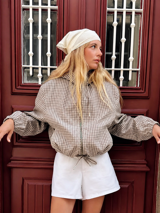 Model in front of a dark red door wears the checked hooded jacket zipped up with white shorts and a cream headscarf. 1