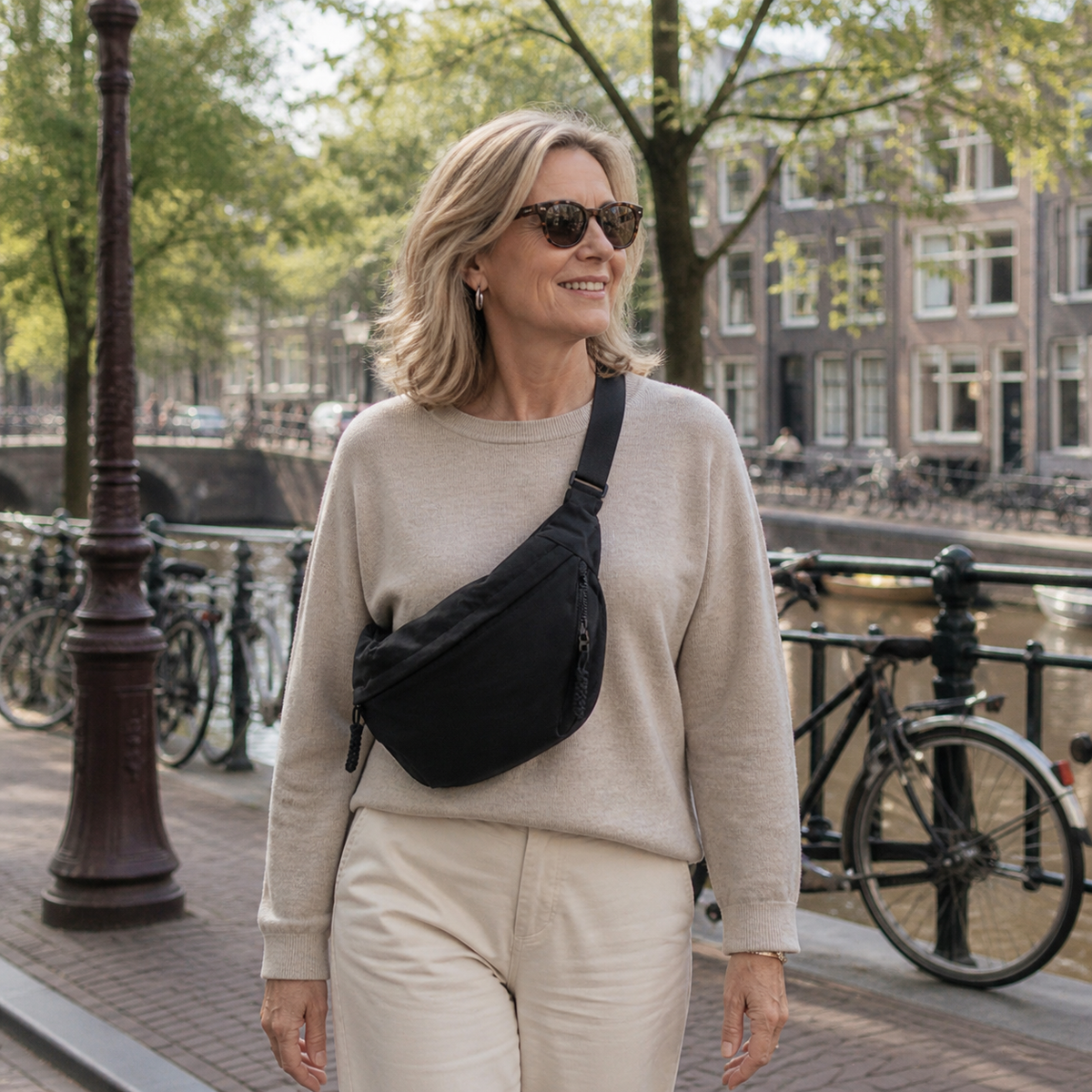 Smiling woman in sunglasses and a cream jumper wears the black bag crossbody while walking along a canal with parked bicycles. 10