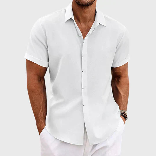 White short-sleeve shirt with rolled cuffs, worn open at the neck by a model in white trousers. 2