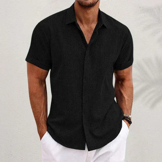 Black short-sleeve shirt with cuffed sleeves worn by a model with hands in the pockets of white trousers. 1