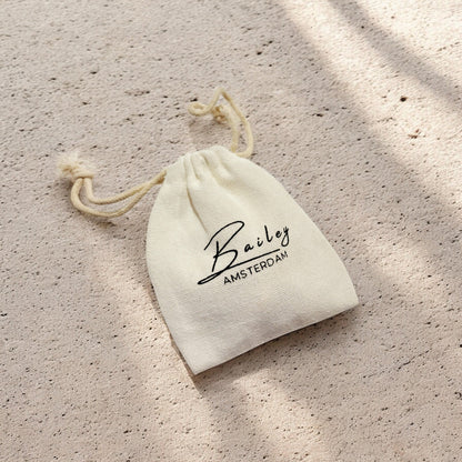 Small cream cotton drawstring pouch with black script lettering, lying on a sandy textured surface in dappled sunlight. 7
