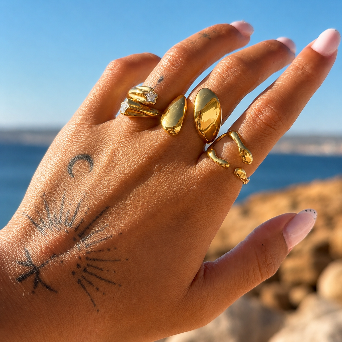 Hand with tattoos wearing the wavy gold-tone open ring alongside other chunky gold rings, with sea and sky behind. 3