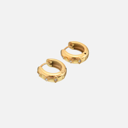 Two gold-tone chunky huggie hoops angled together on a light background, showing their hinged closure and the small clear stones set in the star and eye motifs. 2