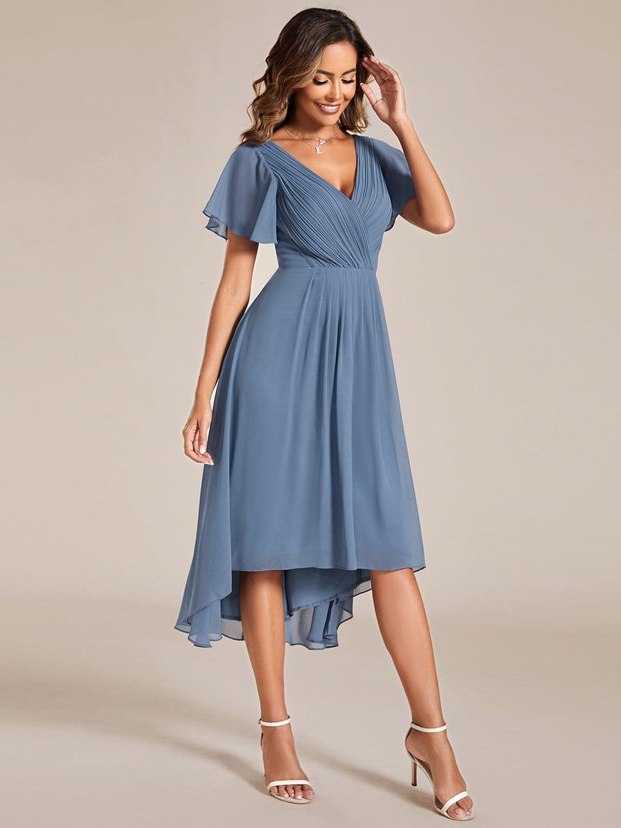 Three-quarter view of the dusty blue dress, model touching her hair, showing the pleated bodice and uneven high-low hem. 9
