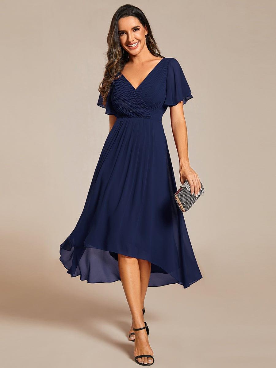 Model in navy chiffon dress with pleated V-neck and flutter sleeves, front view, holding a sparkly clutch and wearing black sandals. 1