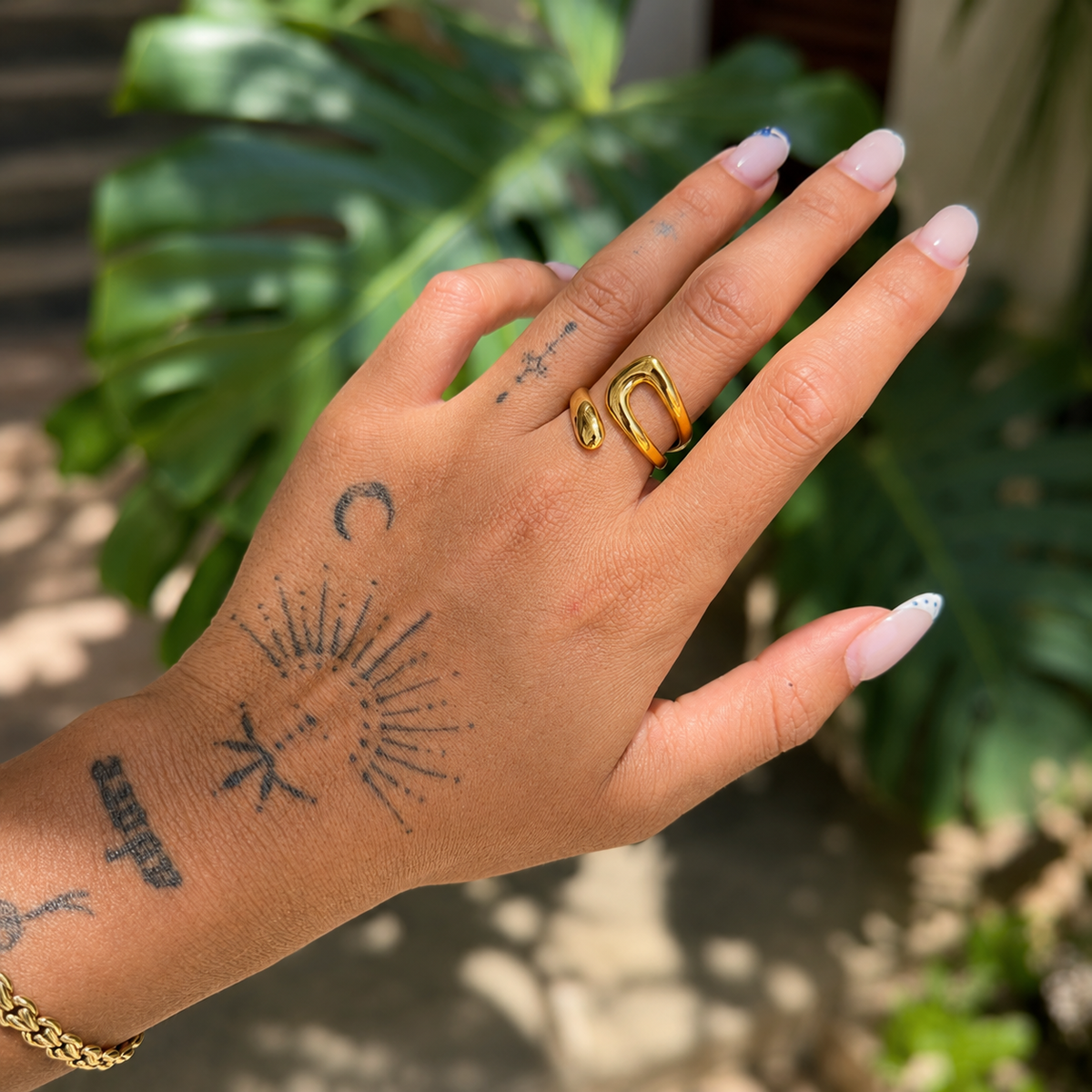 Open gold-tone ring worn on a tattooed hand, with green leaves blurred in the background 3