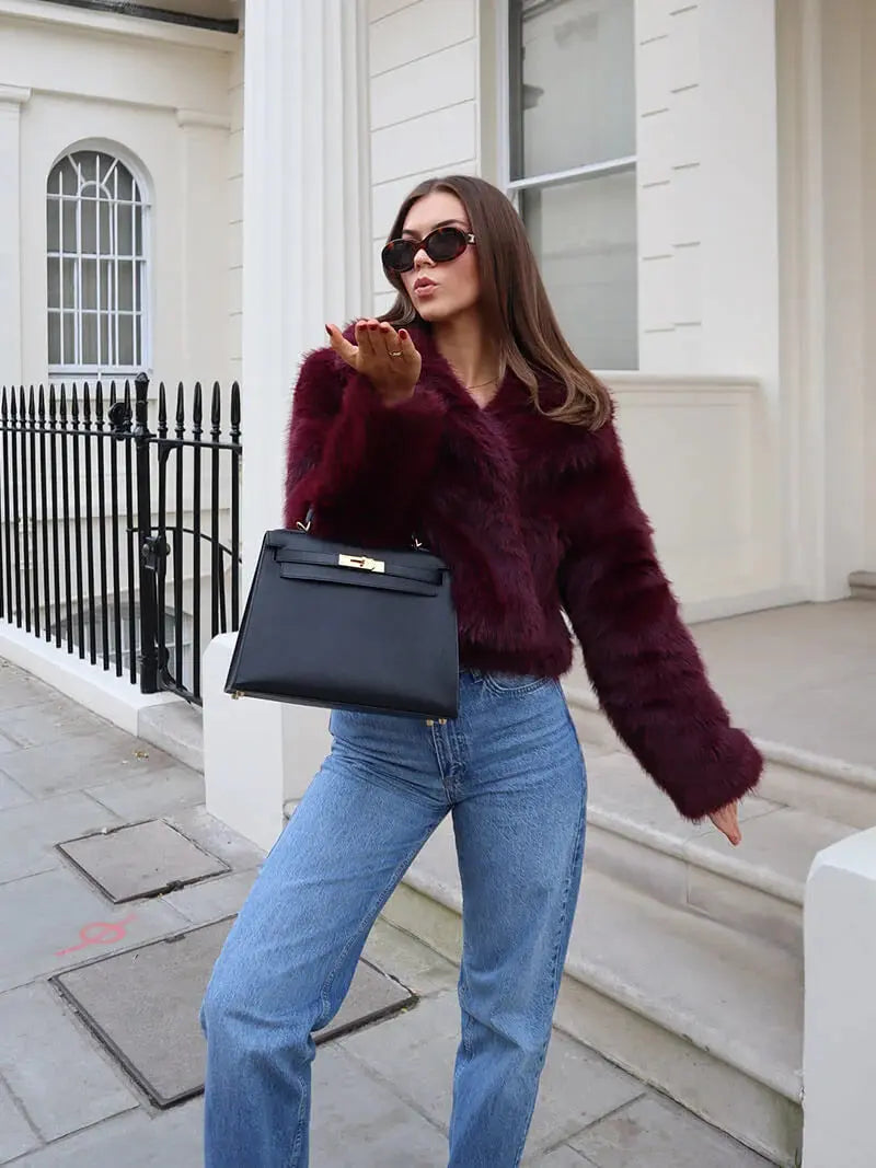 Woman in a burgundy faux fur coat and blue jeans blowing a kiss, holding a black handbag 12
