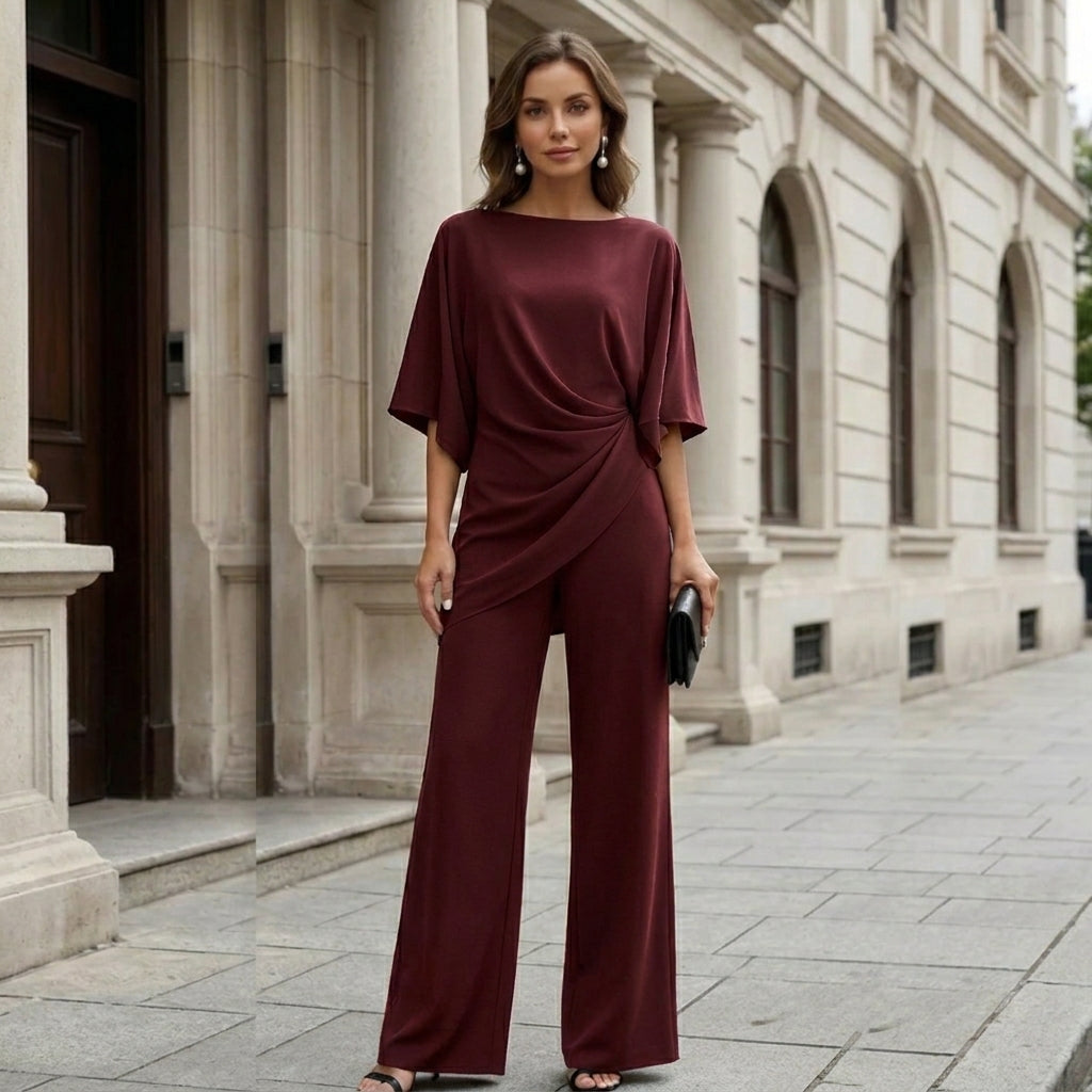 Model standing in front of a stone building in the burgundy set, facing the camera with a black clutch in hand. 11