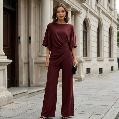 Model standing in front of a stone building in the burgundy set, facing the camera with a black clutch in hand. 11