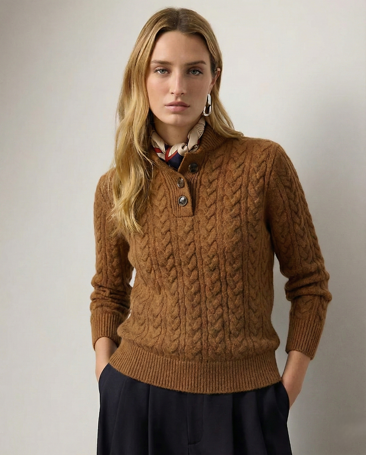 Model facing the camera in a caramel-brown cable-knit sweater with a three-button collar, silk scarf at the neck and dark trousers. 1