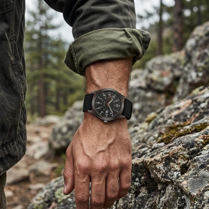 Black-dialled watch with black nylon strap worn on a man's wrist, rolled green sleeve, on a rocky forest trail. 2