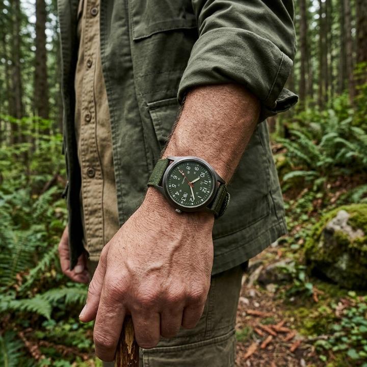 Green-dialled watch with green nylon strap worn on a man's wrist, resting on a walking stick amid ferns in woodland. 3
