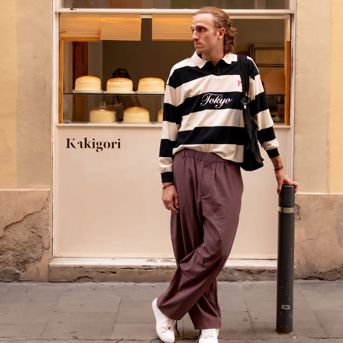 Model in plum-brown elasticated trousers and a striped rugby shirt, leaning on a bollard outside a bakery window. 3