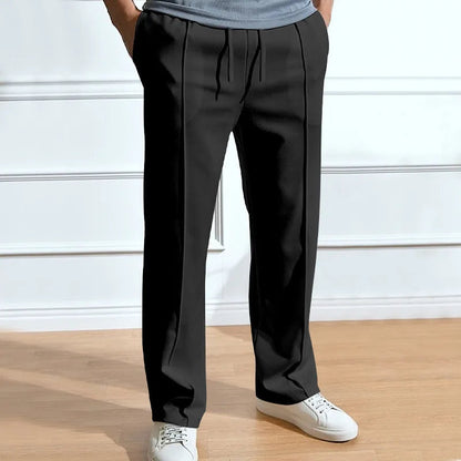 Model wearing the trousers in black, hands in pockets, showing the front creases and drawstring waist against a panelled white wall. 3